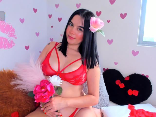 Mia Aylen, con i capelli neri, un fiore di rosa rosa tra i capelli, indossa lingerie di pizzo rossa, tiene in mano fiori rosa e bianchi, è seduta sul letto con cuscini a forma di cuore marroni e neri..
