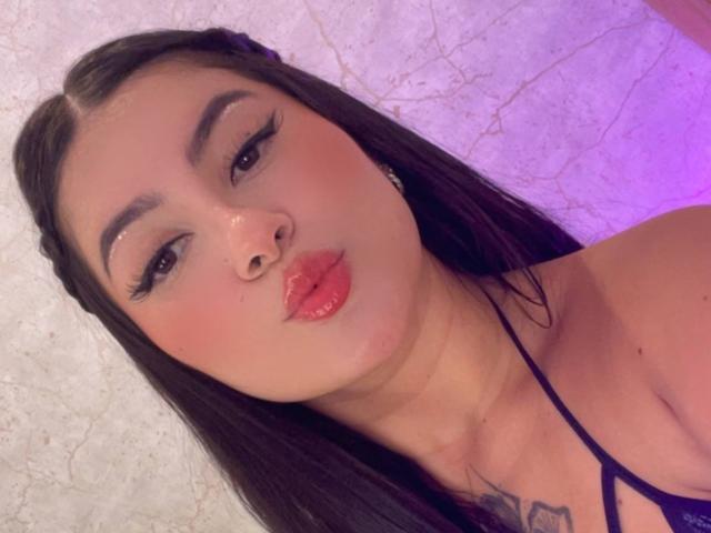 AlanaaWalker with long dark hair, thick eyebrows, and shiny red lips, pouting slightly, wearing a black strappy top, showing a small tattoo on her chest, nose pierced.