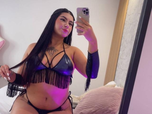 AlanaaWalker with long black hair, wearing a black leather fringe bikini, holding her hair, showing her tattoos, and holding a phone, in a mirror selfie. Her ass is visible.
