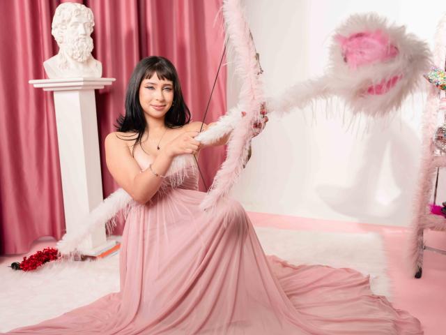 DaliaCastle com cabelo preto, vestido rosa, segurando um arco com penas rosas, sentada em pele branca, busto branco à esquerda, cortinas rosa, flores vermelhas, sorrindo, fofa, sexy, rosa e branco..