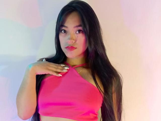 NatalyEscobar with long black hair, pink halter top, and pink lipstick, hand on chest, pink and blue lighting, medium skin, looking at camera, subtle makeup, ring on index finger.