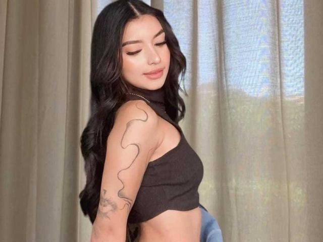 IrisBeaumont with long black wavy hair, wearing a black crop top, showing a wavy tattoo on her right arm, eyes closed, standing in front of beige curtains.