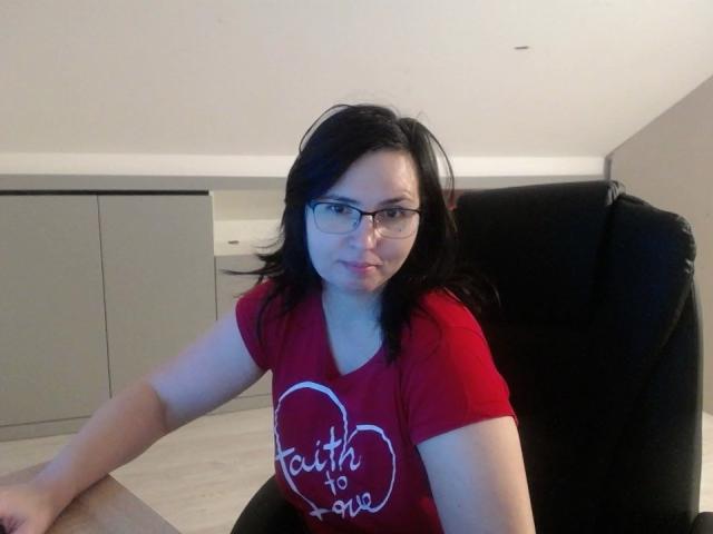 SweetLouise, a fair-skinned woman with black hair and glasses, sits in a black leather chair. She wears a red t-shirt with white text "taught to love" inside a heart. Her expression is neutral, slightly puckered. The background includes a white ceiling and gray cabinets. The image is a casual, straightforward photograph.