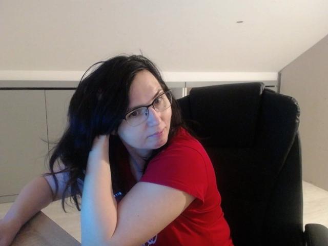 SweetLouise with black hair, glasses, and red shirt, sitting in a black chair, hand in hair, looking slightly bored. Boobs not visible, pussy not visible, ass not visible.