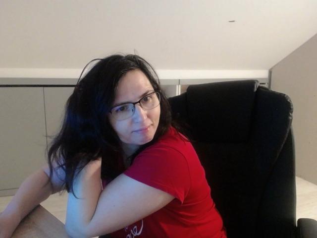 SweetLouise with black hair, glasses, and red shirt, hugging her arm while sitting in a black chair, slightly smiling. Her tits are small, and she has a cute face.