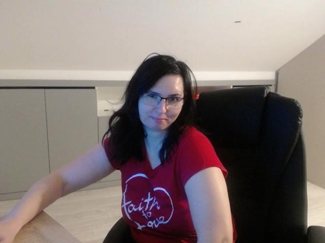 SweetLouise, with glasses and black hair, sits in a black chair, wearing a red shirt with white text, looking at the camera, her ass slightly visible to the side.
