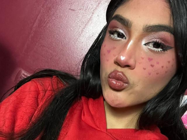 MissSubmissive hat rosafarbene, herzförmige Akne auf ihren Wangen, glänzlich rote Lippen und schwarzen Eyeliner mit dicken Augenbrauen. Sie trägt eine rote Kapuzenjacke und hat langes, schwarzes Haar..