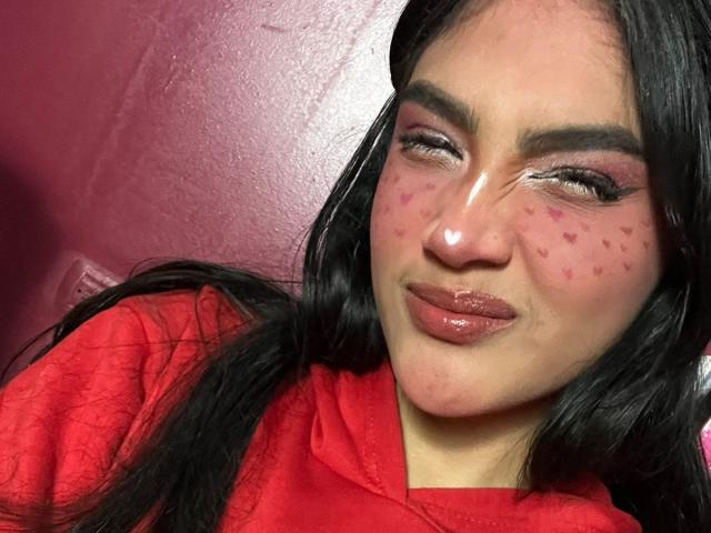 MissSubmissive mit vollen, roten Lippen, schwarzem Haar und rosa Pickelchen auf den Wangen, trägt eine rote Kapuzenjacke und macht ein schmollendes Gesicht in einem Selfie..