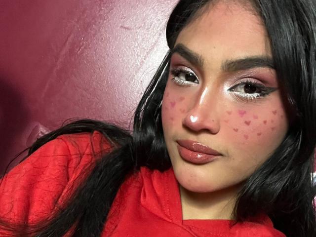 MissSubmissive hat rosafarbene, herzförmige Pickel auf ihren Wangen, glänzende, rosafarbene Lippen und langes, dunkles Haar, trägt eine rote Kapuzenjacke, hat auffälligen Eyeliner und rosafarbenen Lidschatten und blickt in die Kamera..