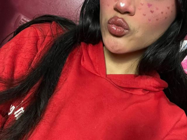 Das Gesicht von MissSubmissive mit glänzenden, rosafarbenen Lippen, schwarzen, welligen Haaren, einer roten Kapuzenjacke und einem rosafarbenen Herzchen-Pickel auf der Wange, die ihre Lippen zu einem Kussmund formt..