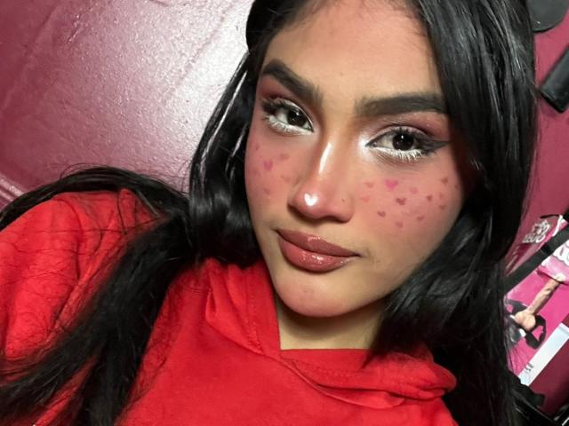 MissSubmissive mit dunklem Haar, roter Kapuzenjacke, rosa Herz-Tattoos auf den Wangen, glänzenden rosa Lippen und weißem Eyeliner, lächelt leicht, in einem rot gestrichenen Raum..