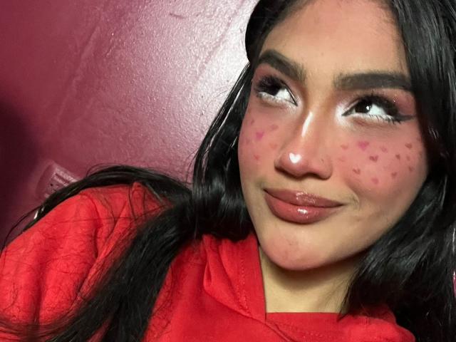 MissSubmissive hat ein süßes Gesicht mit rosa, herzförmigen Pickeln auf ihren Wangen, glänzlichen, rosafarbenen Lippen und dunklem, glattem Haar. Sie trägt eine rote Kapuzenjacke und steht vor einer roten Wand..