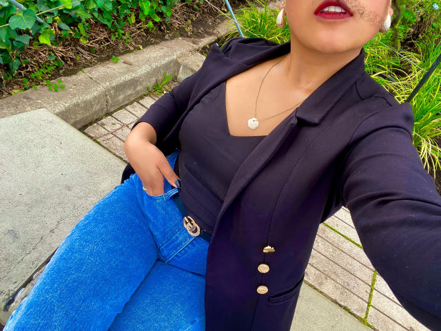 SophieSonyas with dark blazer, blue jeans, black top, red lipstick, and necklace, sitting, hand in pocket, visible tits, medium skin, green background, white earbuds.