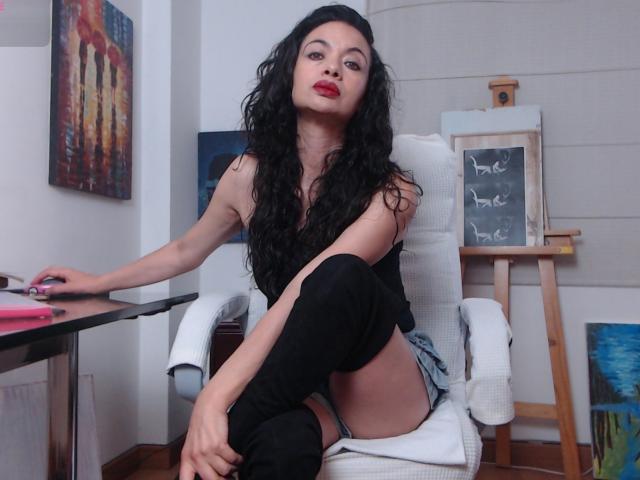 AfroditaRgenis with long black curly hair, red lipstick, black sleeveless top, black thigh-high boots, sitting in white chair, one leg up, revealing her bare ass.