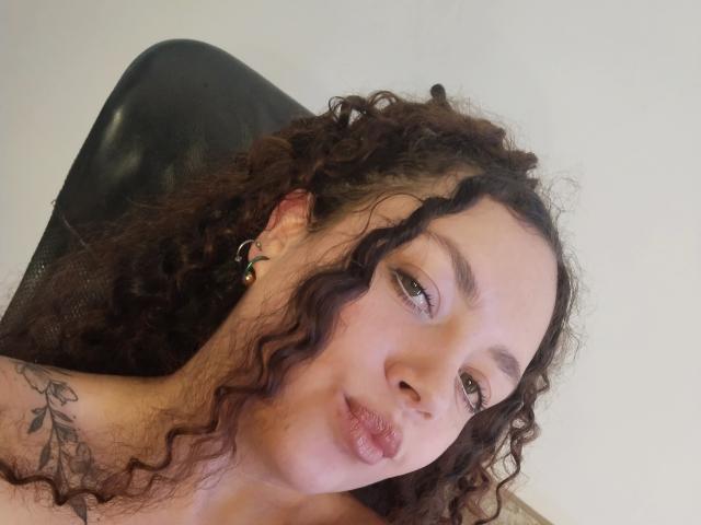 Close-up of RubbyStrongX, a light-skinned woman with curly brown hair and green eyes. She has small, slightly pursed lips and is wearing green earrings. Her right shoulder features a black tattoo with floral designs. The black leather chair she's leaning on contrasts with the white background. Her expression is neutral, and the image has a casual, everyday feel. Her hair is textured with natural curls, and her eyebrows are thin and slightly arched. The photograph is straightforward with a focus on her face and upper shoulder.