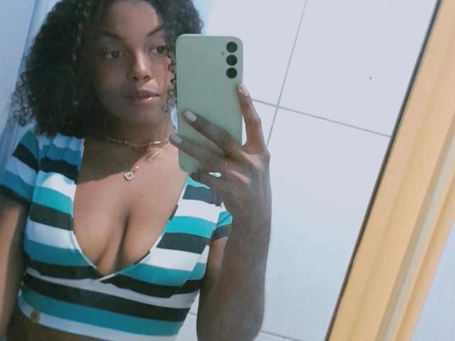 PreatyOne with curly hair, wearing a striped top, showing her tits in a mirror selfie, holding a green phone, necklace, and subtle smile, in a bathroom.