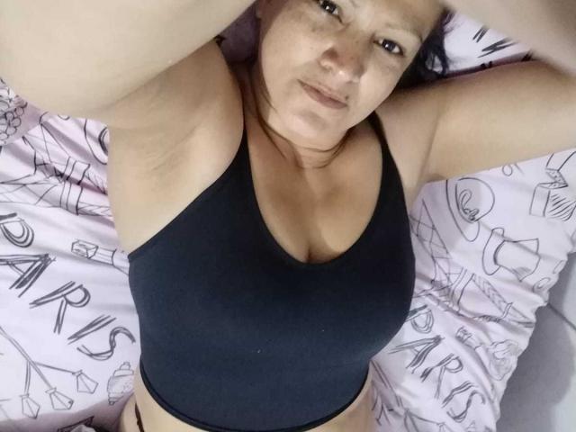 MilenneSexy lying on a bed, wearing a black sports bra, arms raised, showing her armpits and small tits, with a slightly pouty expression on her face.