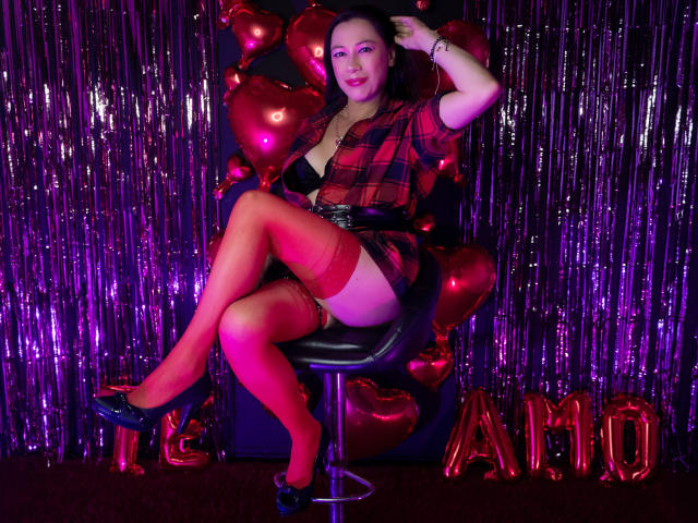 Noemi: Am schönsten mit rotem Karohemd, schwarzem BH, roten Strumpfhosen mit Gürtel, schwarzen High Heels, sitzend auf einem Hocker, umgeben von roten Herzballons und violettem Glitzer, wobei ihre Brüste und ihr Hintern zu sehen sind..