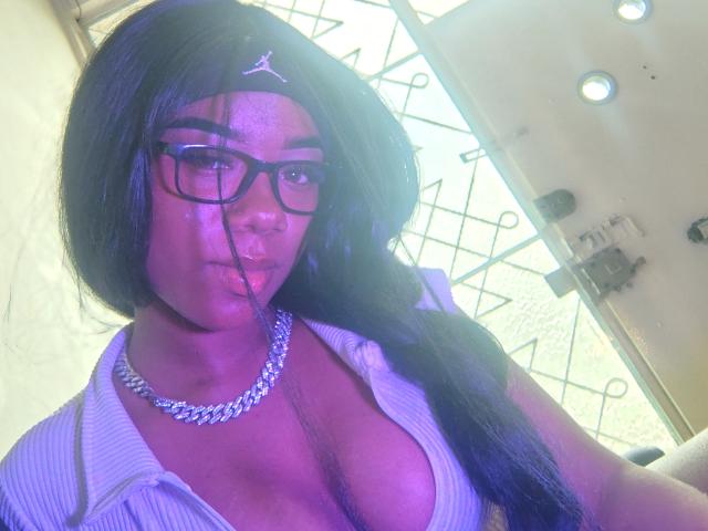 SophiaTaylorn with long black hair, glasses, and a white headband, wearing a low-cut, ribbed shirt, showing big tits, and a silver chain necklace, with purple lighting.