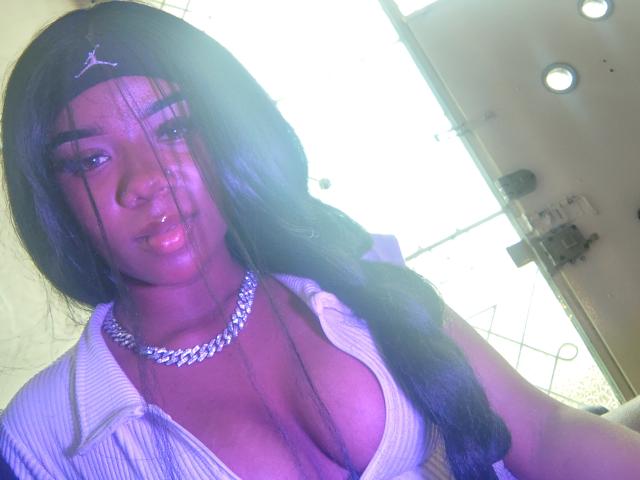 SophiaTaylorn with dark skin, long black hair, a silver chain necklace, and a low-cut white shirt showing her big, shiny tits. Bright sunlight in the background.
