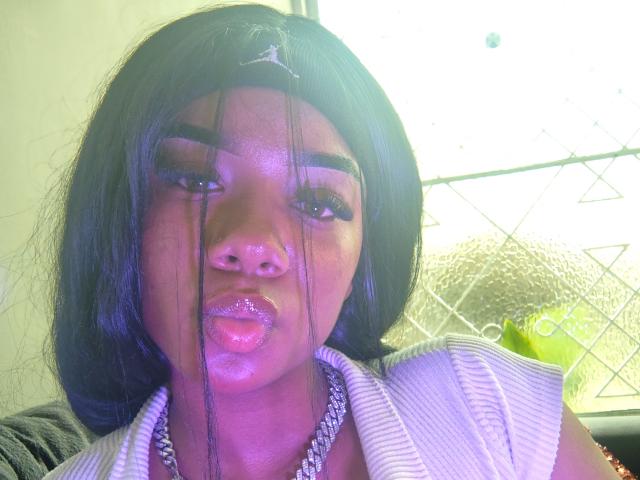 SophiaTaylorn with glossy pink lips, long black hair, and chain necklace, has white liquid dripping from her nose and mouth, wearing a striped shirt, in a bright photo.