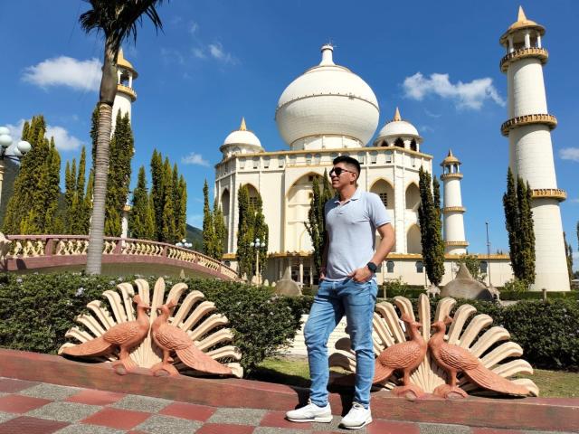 SanderNoah stands in front of a white, domed, and towered building with peacock statues, wearing a grey t-shirt, blue jeans, white sneakers, and sunglasses, under a clear blue sky.