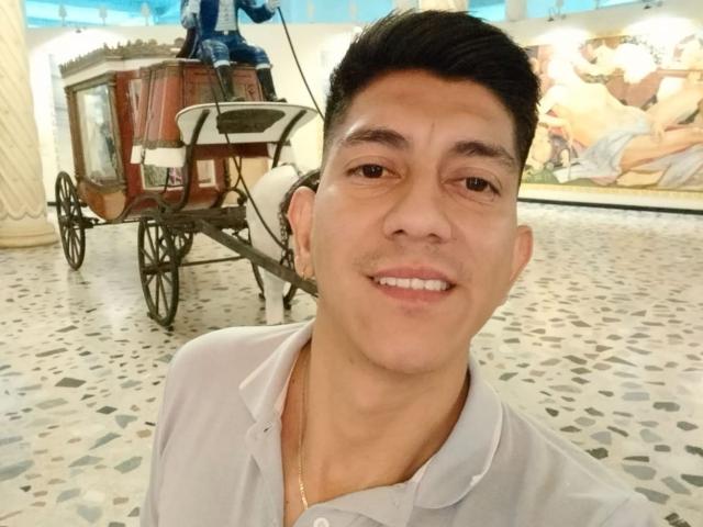 SanderNoah has a medium skin tone, short black hair, and a slight mustache, wearing a white shirt, smiling with a gold necklace, in front of a wooden cart.