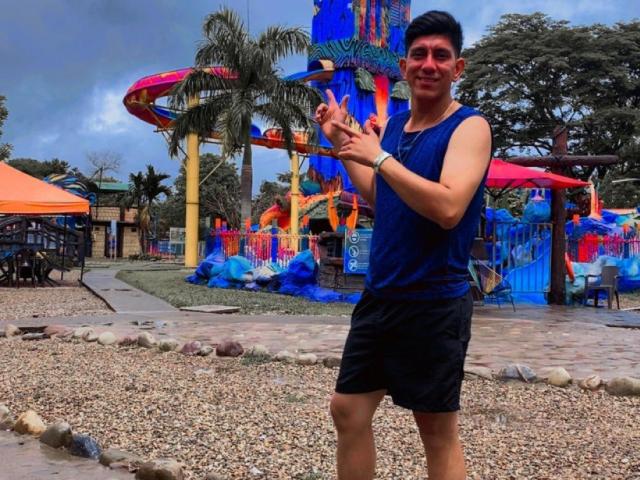 SanderNoah stands in a pebble-covered area, wearing a blue sleeveless shirt and black shorts, holding a colorful water gun, smiling with short dark hair, in a water park.