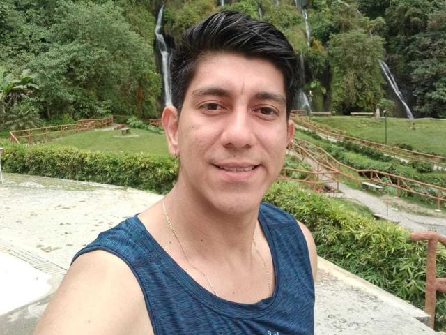 SanderNoah with short black hair, wearing a blue tank top, smiling, has a slight tan, and is in a park with a waterfall. His ass is not visible.
