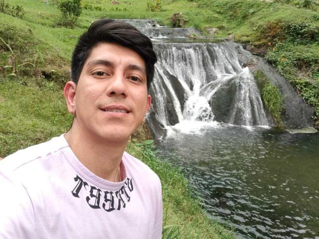 SanderNoah with short black hair, light skin, and a mustache, wearing a pink shirt, standing in front of a waterfall. Water's white, green grass surrounds.