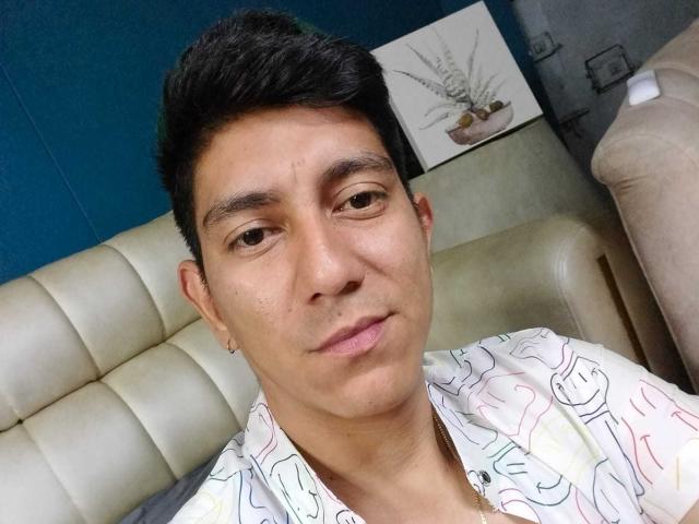 SanderNoah with short black hair, medium brown skin, wearing a patterned shirt, small earrings, and a neutral expression, sitting on a beige leather couch.
