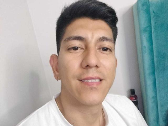 SanderNoah has short black hair, brown eyes, and a slight smile. He's wearing a white shirt, has a small earring, and a gold chain. His nose is small.