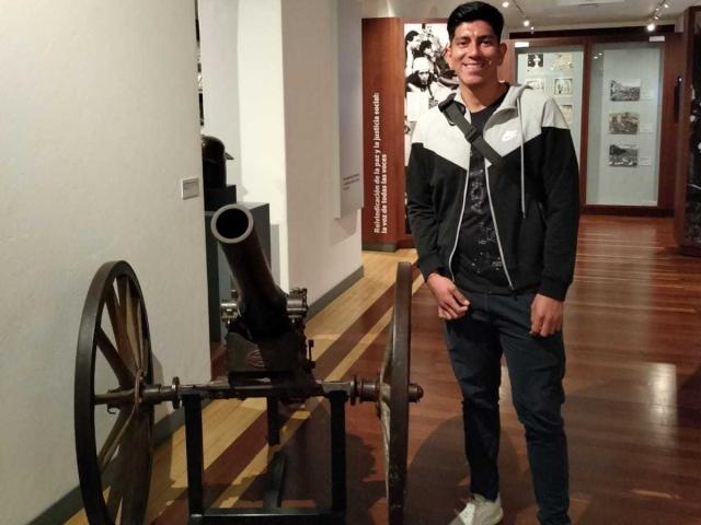 SanderNoah stands in a museum, wearing a black and white hoodie, dark jeans, and white sneakers, next to a old cannon with wooden wheels on a wooden floor.