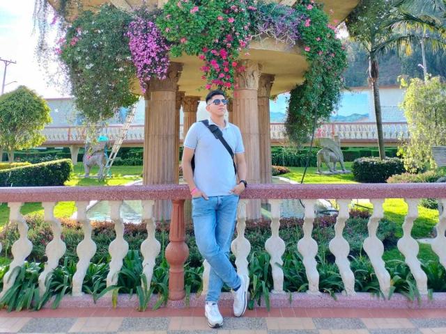 SanderNoah stands against a pink railing, wearing a gray t-shirt, blue jeans, white sneakers, black strap, and sunglasses, with pink and purple flowers overhead.