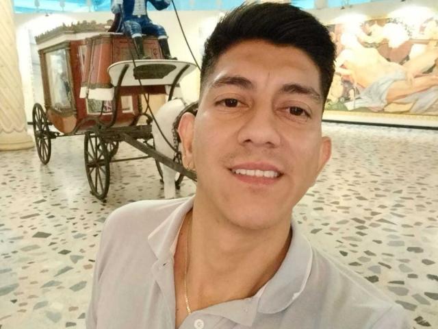 SanderNoah, a cute dude with short black hair, wearing a white polo, smiling at the camera, standing in front of a wooden cart with wheels in a tiled room.