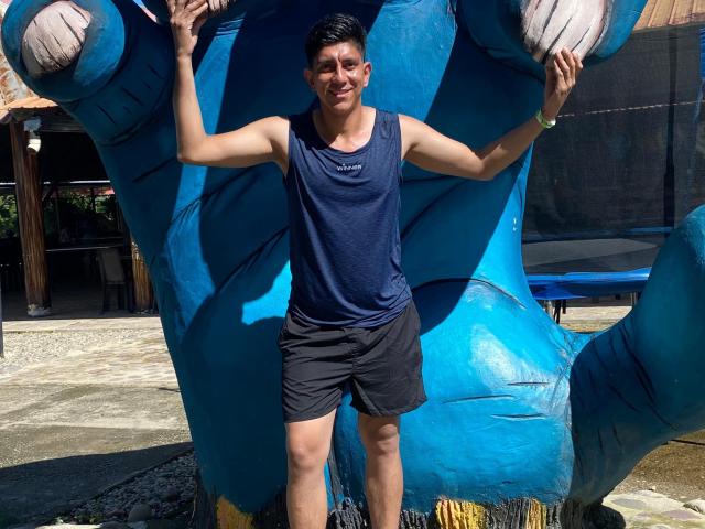 SanderNoah stands in front of a blue inflatable tiger, arms raised, wearing a dark blue tank top and black shorts, smiling with short dark hair and a tan complexion.