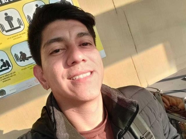 SanderNoah, with short black hair and a slight smile, wearing a dark jacket over a brown shirt, has a backpack strap and a small earring on his left ear.