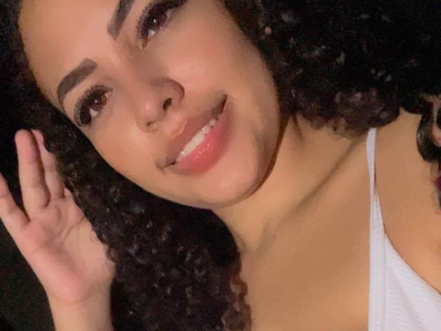 Geyza with curly black hair, brown eyes, and a smile, wearing a white tank top, showing a cute face with a slight nose and full lips. She's touching her hair.
