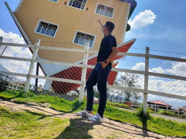 SanderNoah stands in front of a tilted yellow building, pointing upwards with his right hand. He's wearing a black t-shirt, blue jeans, white Adidas sneakers, and a black cap.