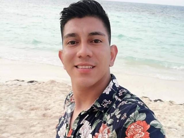 SanderNoah with short black hair, wearing a floral shirt, smiling at the camera on a sandy beach with turquoise water in the background. Slightly chubby, tan skin.