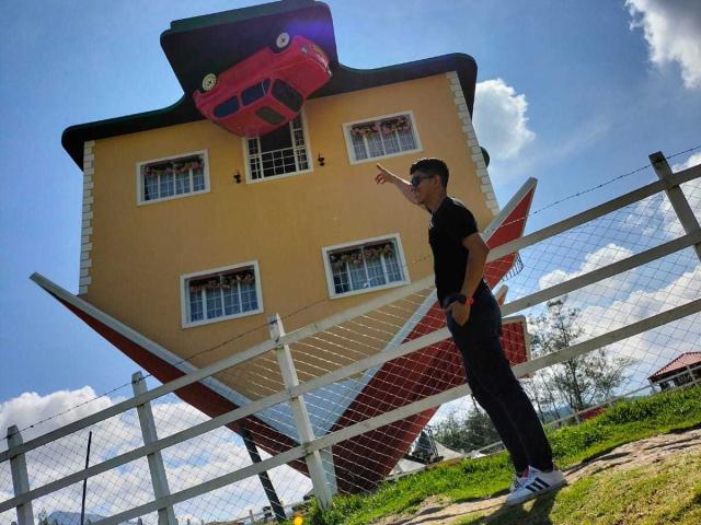 SanderNoah stands in front of a tilted house with a red car on the roof, wearing a black t-shirt and black pants, pointing up at the car.