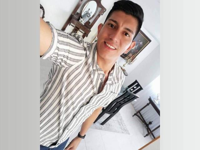 SanderNoah with short black hair, smiling, wearing a striped shirt, standing in a room with a wooden mirror and black furniture, taking a selfie.
