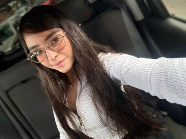 EvaMcAllister with long brown hair, wearing glasses and a white ribbed shirt, taking a selfie in a car, showing a slight smile and her small tits.