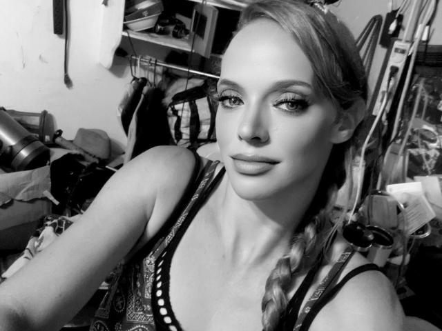 SuggerX has a sexy face, pouty lips, and small tits in a black-and-white photo with braided hair, wearing a patterned top in a cluttered room.