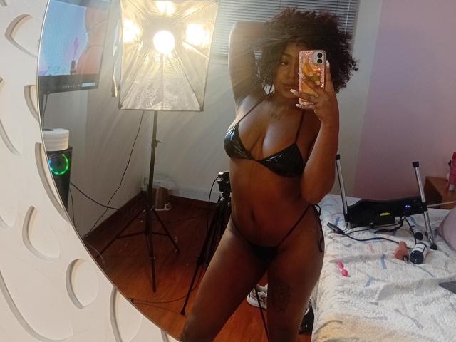 AmberQueeny in black bikini, showing off her tits and ass, with a camera flash in the mirror, holding a phone with a colorful case, in a bedroom.