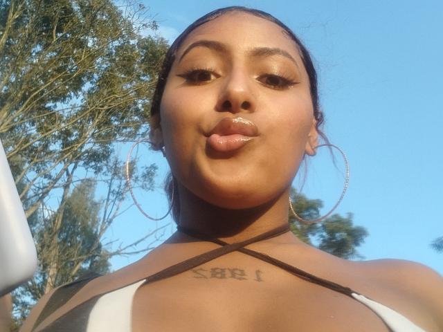 AmberQueeny has glossy lips, hoop earrings, and a tattoo on her chest. She's wearing a black and white bikini top, looking down at the camera.