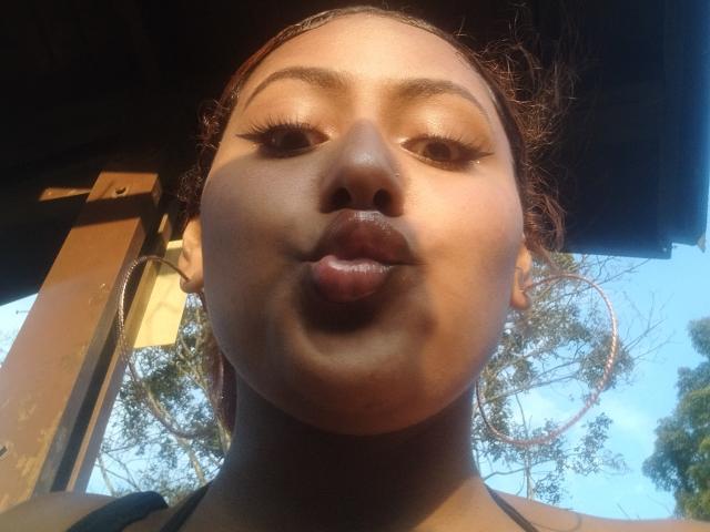 AmberQueeny's puffy lips are centered in the shot, her brown eyes looking forward, with her dark brown skin glowing in sunlight. She wears large hoop earrings.