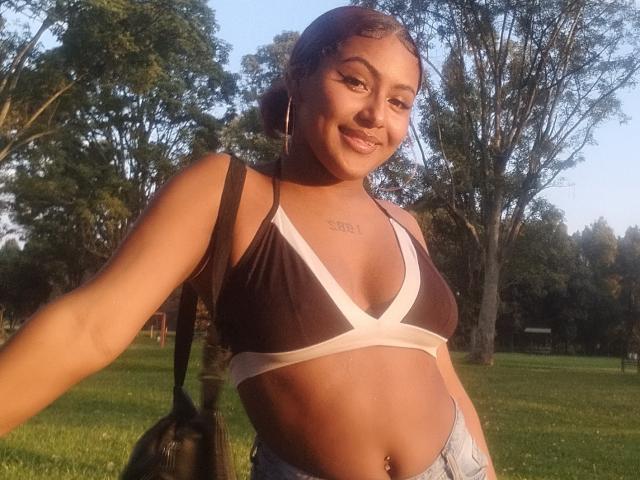 AmberQueeny with medium brown skin, curly black hair, wearing a brown and white bikini top, showing her small tits, smiling, with a black bag on her shoulder.