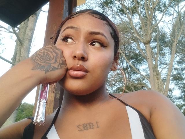 AmberQueeny with dark skin, braids, and a rose tattoo on her hand, pouting with her nose ring, wearing a white and black top, hoop earrings, and a necklace.