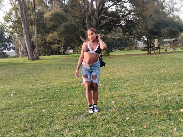 AmberQueeny with medium tits, wearing a white bra and ripped denim shorts, standing in a grassy park, holding a phone to her ear, in black and white sneakers.